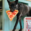 Load image into Gallery viewer, P.L.A.Y. Snack Attack Collection - Puppy-roni Pizza Toy in beautiful black dog's mouth coming around kitchen island with pizza box on ground
