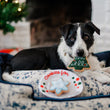 Load image into Gallery viewer, P.L.A.Y. Merry Woofmas Christmas Eve Cookies - black and white dog with Christmas Tree cookie in it's mouth laying in a lounge bed with Christmas Tree and fireplace in the background