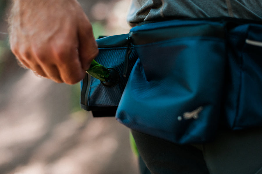 P.L.A.Y.'s Explorer Pack in Waterfall Blue with hand pulling poop bags out of the dispenser