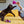 Load image into Gallery viewer, P.L.A.Y. Feline Frenzy Fig N’ Whiskers Toy Set  - A person dangling the fig slice toy with bright pink feathers bursting out of the top above a beautiful cat&#39;s head, while the cat tries to paw it, on a yellow bed.
