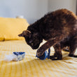 Load image into Gallery viewer, A black and brown cat playing with P.L.A.Y. Feline Frenzy Kitty Crumble Toy Set on a yellow bedspread
