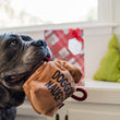 Load image into Gallery viewer, P.L.A.Y. Home for the Holidays Collection Fur-Ever Naughty - big black dog holding one side of the bag of coal in his mouth looking to the side standing in front of a bench with a gift bag in the background and Christmas Tree green pillows
